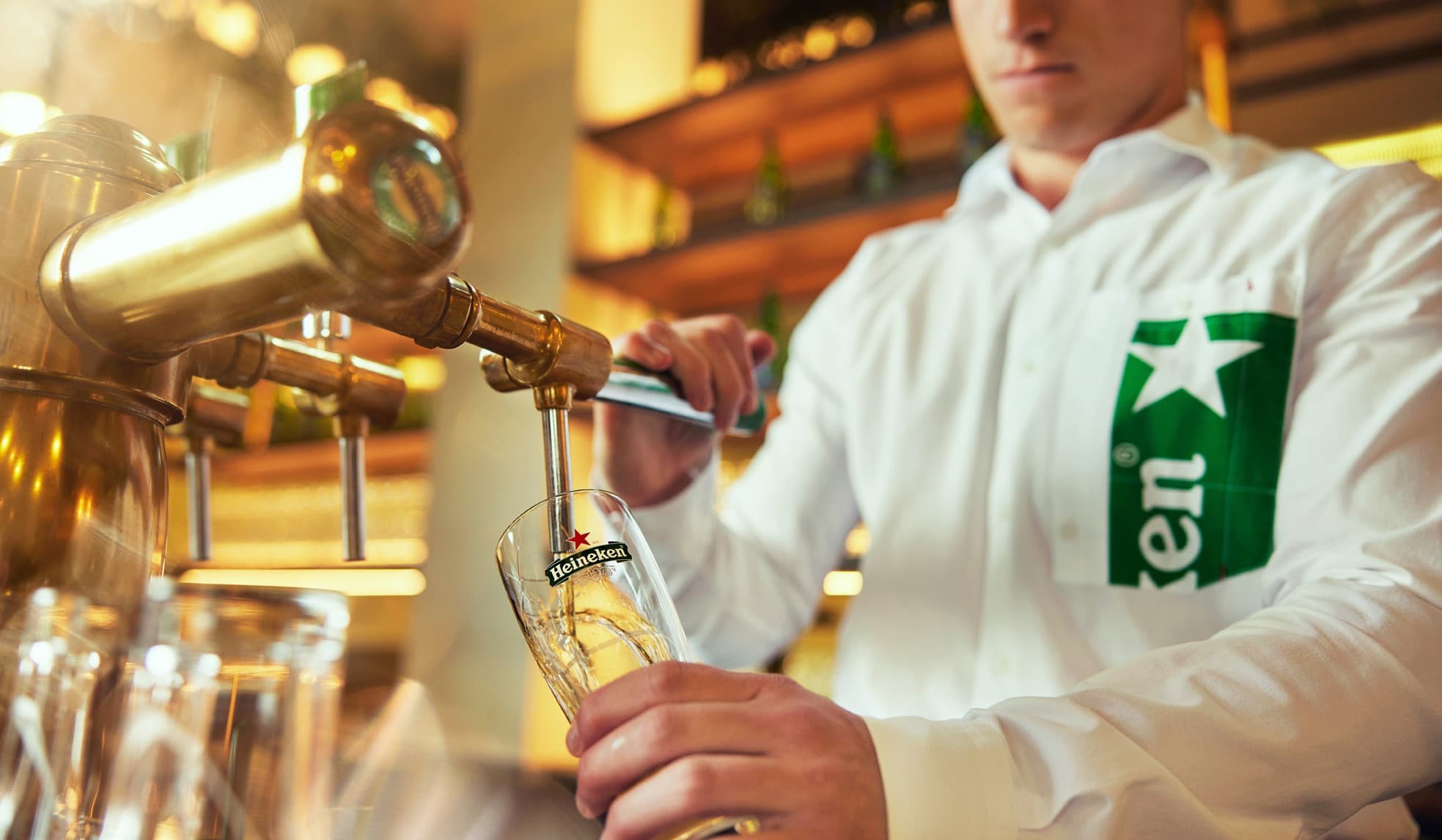 A bartender pours a draft Heineken beer into a branded glass from a brass tap at a bar, wearing a white shirt with a large green Heineken logo on the sleeve.